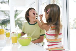 Girl (8-10) at table, feeding brother (8-10) cereal